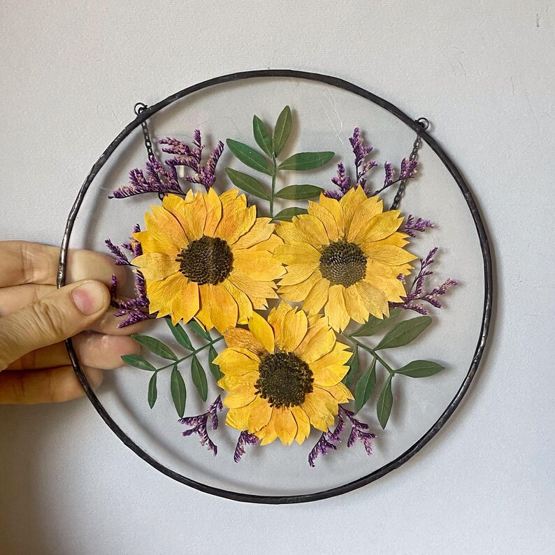Op de afbeelding: Ronde, heldere glazen wanddecoratie met drie gele zonnebloemen met donkerbruine centra. Groene bladeren en paarse takjes omringen de bloemen, alles in een zwart metalen frame. Een ketting is bevestigd om op te hangen.