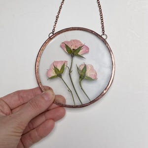May include: A circular glass wall hanging framed in copper, featuring three pressed pink rose blossoms with green stems and leaves. The piece is suspended by a matching copper-toned metal chain attached to the top of the circular frame.