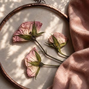 May include: A circular wall hanging featuring three pressed pink roses with green stems and sepals encased within a hammered copper frame. The piece is suspended by a thin metal chain and displays delicate, dried floral details in soft hues.