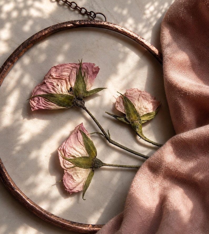 May include: A circular wall hanging featuring three pressed pink roses with green stems and sepals encased within a hammered copper frame. The piece is suspended by a thin metal chain and displays delicate, dried floral details in soft hues.