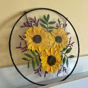 Op de afbeelding: Een ronde, heldere glazen wanddecoratie met drie gele zonnebloemen in het midden. De bloemen hebben donkerbruine centra en zijn omgeven door groene bladeren en paarse takjes, alles in een zwart metalen frame. Een ketting is bevestigd om op te hangen.