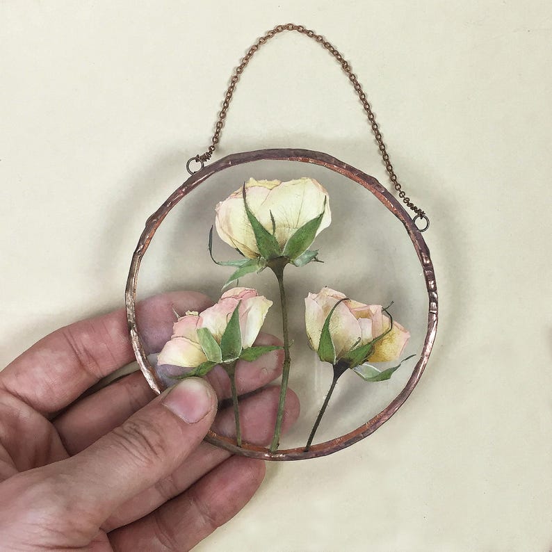 May include: A circular wall hanging featuring three pressed, light-colored roses encased between two clear glass panes. The glass is framed in a textured, dark copper-toned metal border and includes a matching copper-colored metal chain for hanging.