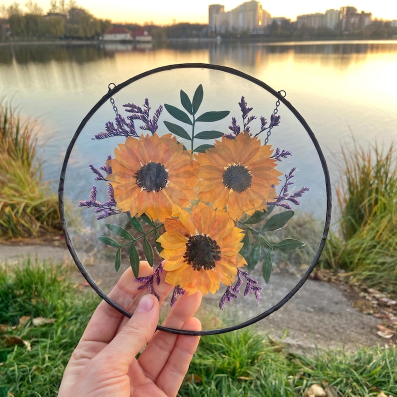 Op de afbeelding: Een ronde glazen plaat met een zwart metalen frame, met geperste zonnebloemen en groen. De zonnebloemen zijn helder geel-oranje met donkere centra, aangevuld met paarse en groene accenten. Een decoratief bloemstuk.