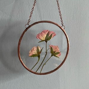 May include: A circular wall hanging featuring three pressed pink roses encased between two clear glass panes. The glass is framed by a hammered copper border and suspended by a matching copper chain attached to two small rings at the top.