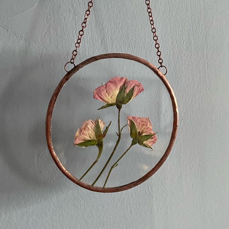May include: A circular wall hanging featuring three pressed pink roses encased between two clear glass panes. The glass is framed by a hammered copper border and suspended by a matching copper chain attached to two small rings at the top.