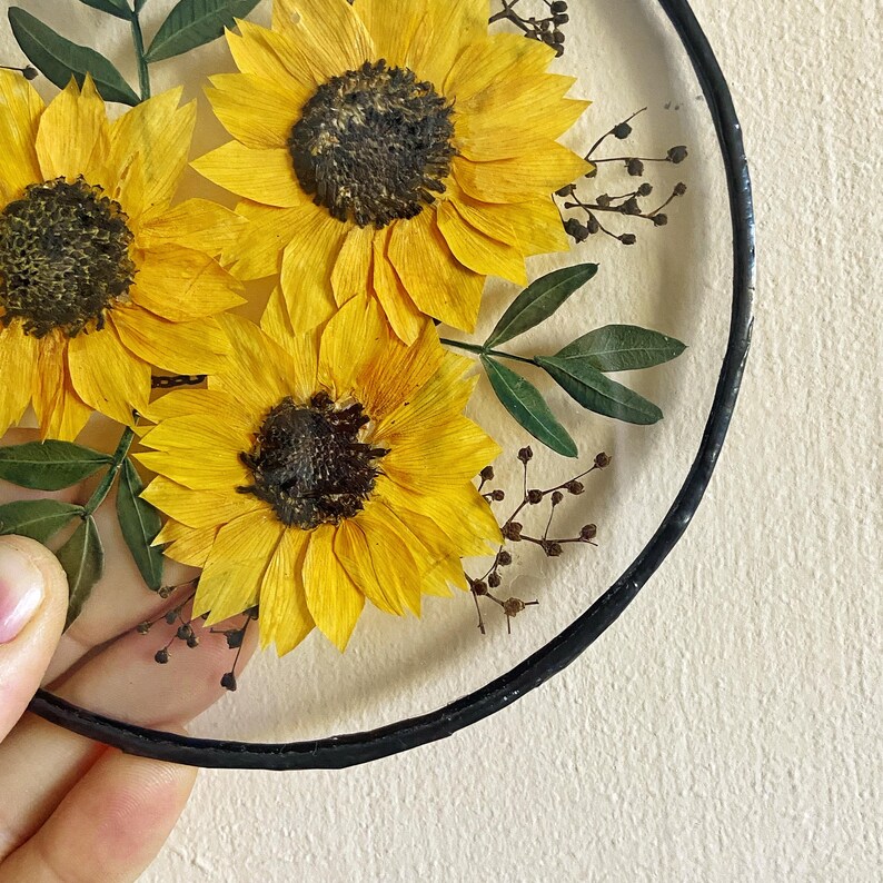 Op de afbeelding: Een helder, rond decoratief stuk met drie felgele zonnebloemen met donkerbruine centra. Groene bladeren en kleine bruine takjes zijn ook zichtbaar. Het stuk is omlijst met een zwarte rand.