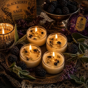 May include: Four round soy candles in clear glass containers, each with a lit wick, scattered dried herbs, and dark stones on top. The candles are arranged on a dark metal tray with fresh sage leaves and blackberries, near a purple bottle.
