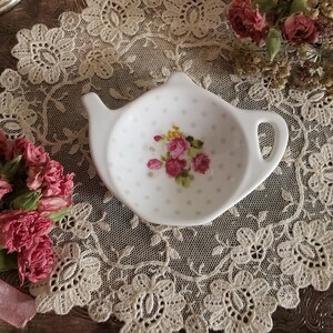 May include: White ceramic teapot-shaped tea bag holder with a floral design of pink roses and yellow accents. The holder has a handle and spout, and is decorated with small grey polka dots. A charming addition to any tea set.