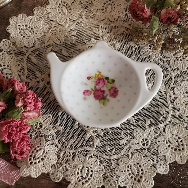 May include: White ceramic teapot-shaped tea bag holder with a floral design of pink roses and yellow accents. The holder has a handle and spout, and is decorated with small grey polka dots. A charming addition to any tea set.