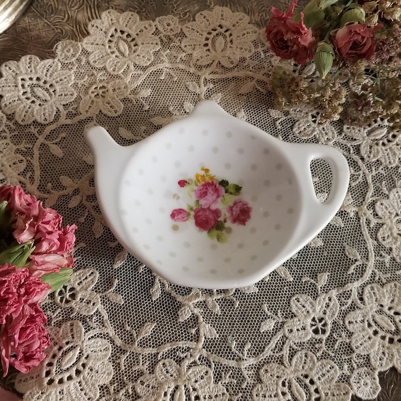 May include: A white ceramic teapot-shaped tea bag holder with a handle and spout. It has a pattern of small grey polka dots and a floral bouquet of pink and yellow roses. The background is a lace doily.
