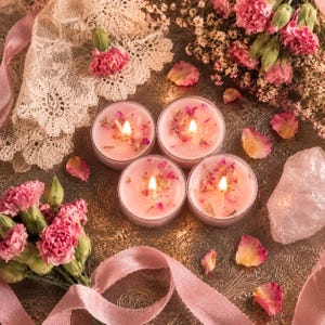 May include: Four round pink candles in glass containers with lit wicks, decorated with dried flower petals. The candles are arranged on a metallic tray alongside pink carnations, white lace fabric, a raw rose quartz crystal, and pink ribbon.