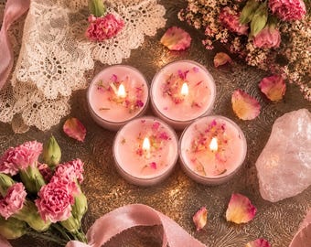 May include: Four round pink candles in glass containers with lit wicks, decorated with dried flower petals. The candles are arranged on a metallic tray alongside pink carnations, white lace fabric, a raw rose quartz crystal, and pink ribbon.