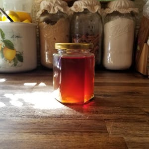 May include: A small, hexagonal glass jar filled with a reddish-brown liquid, possibly honey or syrup. The jar has a gold-colored lid and sits on a wooden surface. Other jars with various contents are visible in the background.