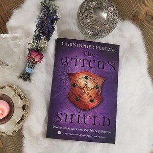May include: A book titled "The Witch's Shield" by Christopher Penczak, featuring a shield graphic on the cover. The book is on a white fluffy surface, with a bundle of dried flowers and a silver container nearby.