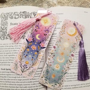 May include: Two decorative bookmarks with celestial designs. Each bookmark features a gradient background with moons, stars, and floral accents. One has a pink tassel, the other a purple tassel. The bookmarks are on a book page.