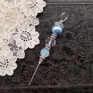 May include: A decorative hat pin with a silver-tone metal needle and a beaded top. The top features a large, pearlescent blue bead, clear crystal beads, and a smaller light blue bead. The pin is set against a dark, textured background with white lace.
