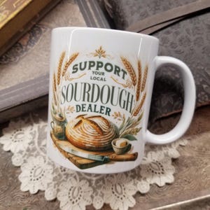 May include: White ceramic mug with the text "SUPPORT YOUR LOCAL SOURDOUGH DEALER" in green and brown. The mug features illustrations of wheat, a loaf of bread, a knife, and butter.