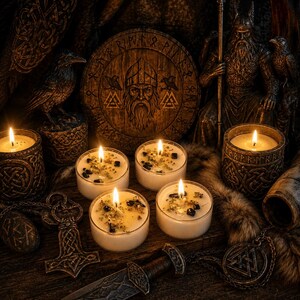 May include: Four small white candles in clear glass containers, each topped with dark stones and dried herbs. The scene includes a circular wooden plaque with a bearded figure, a metal hammer pendant, a dagger, and a statue of a seated warrior.