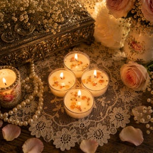 May include: Four small round glass candles with lit wicks, each topped with dried flower petals and amber-colored resin pieces. The candles are arranged on a white lace doily alongside a string of pearls, a floral vase, and a metal box.