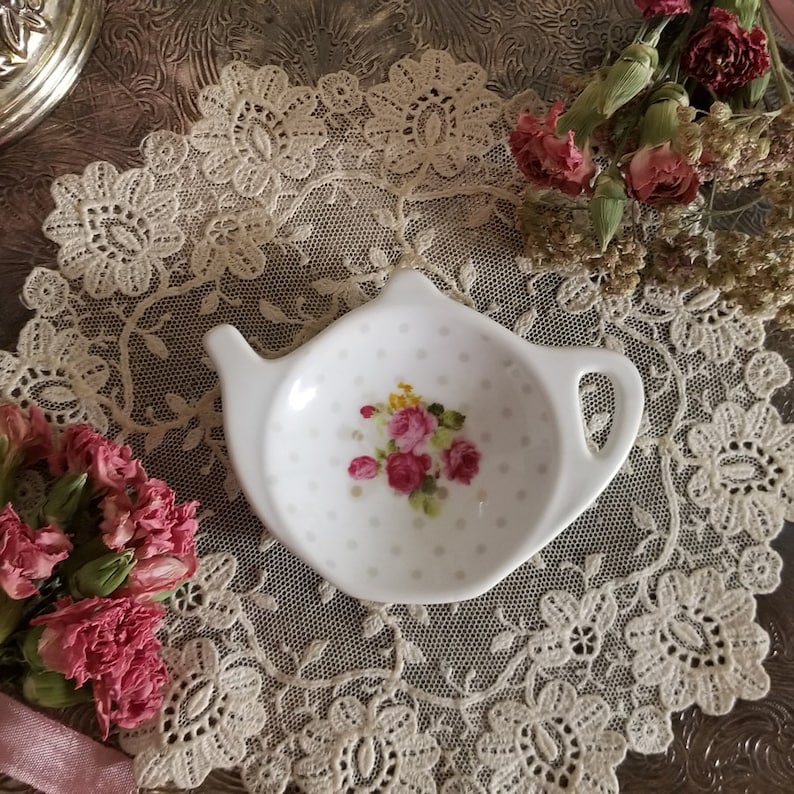 May include: A white ceramic teapot-shaped tea bag holder with a floral design. The holder has a polka dot pattern and a small bouquet of pink and yellow roses. The holder is on a lace doily.