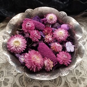 May include: A silver-colored, scalloped bowl filled with dried flowers. The flowers are primarily pink and purple, with a few yellow centers. The bowl has a decorative edge and sits on a lace surface.