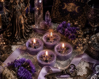 Könnte beinhalten: Vier kleine runde violette Kerzen in Glasbehältern mit brennenden Dochten, verziert mit getrockneten Blumen und Goldflocken. Die Kerzen stehen auf einer weißen Spitzendecke neben einer Bronzestatue der Hekate, einem Schlüssel und einem Buch namens HECATE.