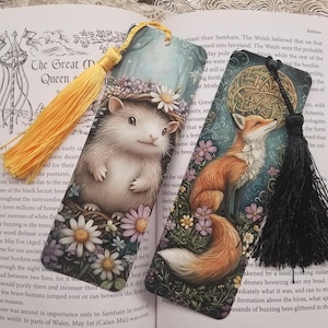 Könnte beinhalten: Zwei dekorative Lesezeichen mit Quasten. Eines zeigt einen Igel mit einem Blumenkranz, umgeben von Blumen. Das andere zeigt einen Fuchs unter einem Vollmond, umgeben von Blumen. Beide Lesezeichen befinden sich auf einem Buch.