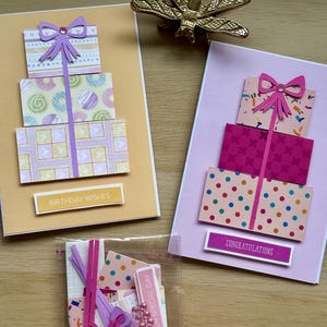May include: Handmade greeting cards with gift box designs. One card says "Birthday Wishes" and another says "Congratulations". The cards feature pink, purple, and yellow colours.