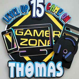 May include: A layered paper cake topper featuring a black diamond shape with yellow borders, the text "GAMER ZONE," and the number "15." It includes cutouts of a smartphone, a computer monitor, and headphones, with the name "THOMAS" at the bottom.
