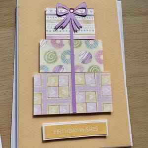 May include: A handmade birthday card featuring a three-tiered gift design. Each tier is decorated with patterned paper in pastel colors. A purple bow tops the design, and the words "BIRTHDAY WISHES" are printed on a rectangular label.