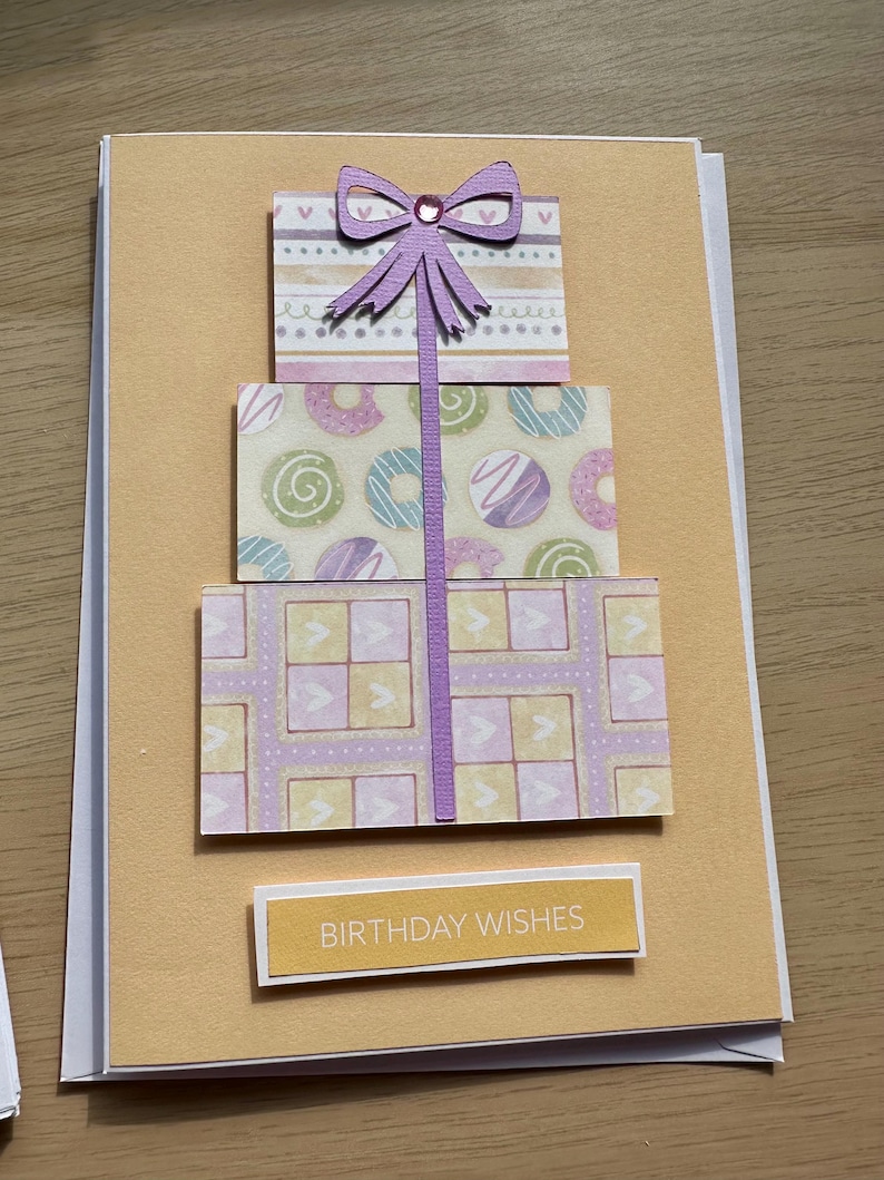 May include: A handmade birthday card featuring a three-tiered gift design. Each tier is decorated with patterned paper in pastel colors. A purple bow tops the design, and the words "BIRTHDAY WISHES" are printed on a rectangular label.