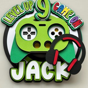 May include: A layered paper cake topper featuring a green game controller design, the number 9, and the name "Jack". The words "Level Up" and "Game On" are included. A black headset with red accents is on the right.
