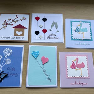 May include: A selection of handmade greeting cards. Designs include a house, champagne glasses, a stork, a butterfly, and heart-shaped balloons. Text includes "Happy New Home", "Happy Anniversary", "baby", "With Sympathy", and "thank you".