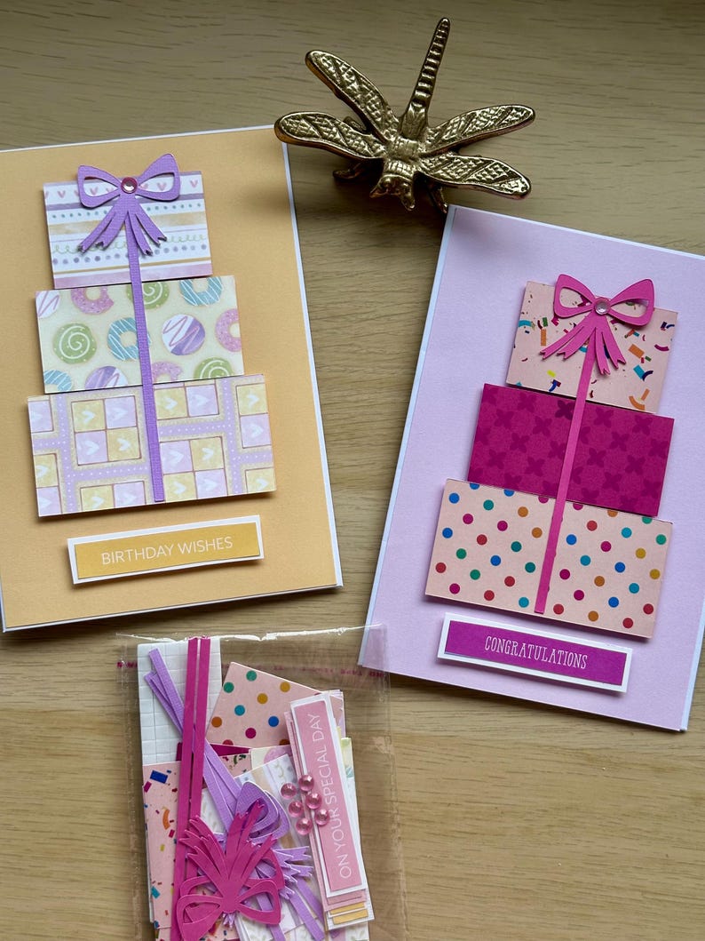 May include: Two handmade greeting cards with gift box designs. One card is yellow with "Birthday Wishes" text, the other is pink with "Congratulations" text. A gold dragonfly figurine sits above the cards. A small bag of craft supplies is also visible.