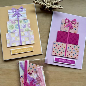 May include: Handmade greeting cards with gift box designs. One card has "Birthday Wishes" text, another says "Congratulations". The cards feature layered paper with bows and colorful patterns. A small bag contains additional crafting supplies.