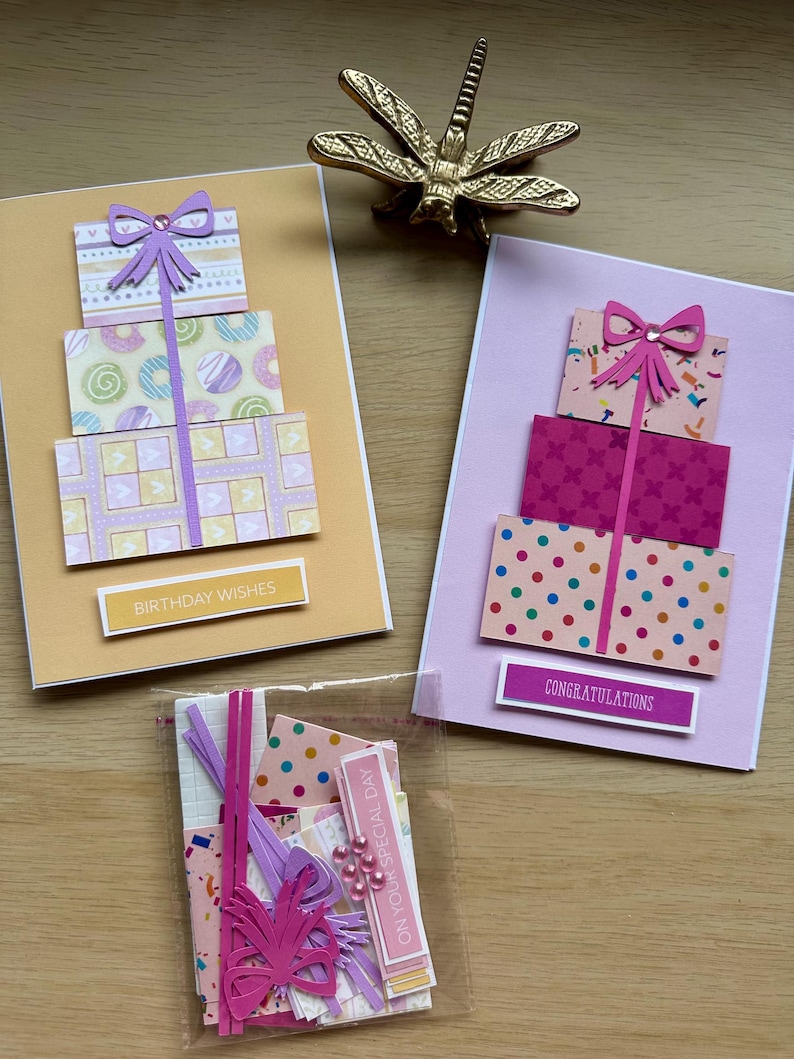 May include: Handmade greeting cards with gift box designs. One card has "Birthday Wishes" text, another says "Congratulations". The cards feature layered paper with bows and colorful patterns. A small bag contains additional crafting supplies.