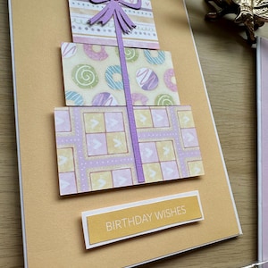 May include: A handmade birthday card featuring three stacked gift boxes with patterned paper. The top box has a purple bow. The middle box has donut designs. The bottom box has a geometric pattern. The card includes the text "BIRTHDAY WISHES".