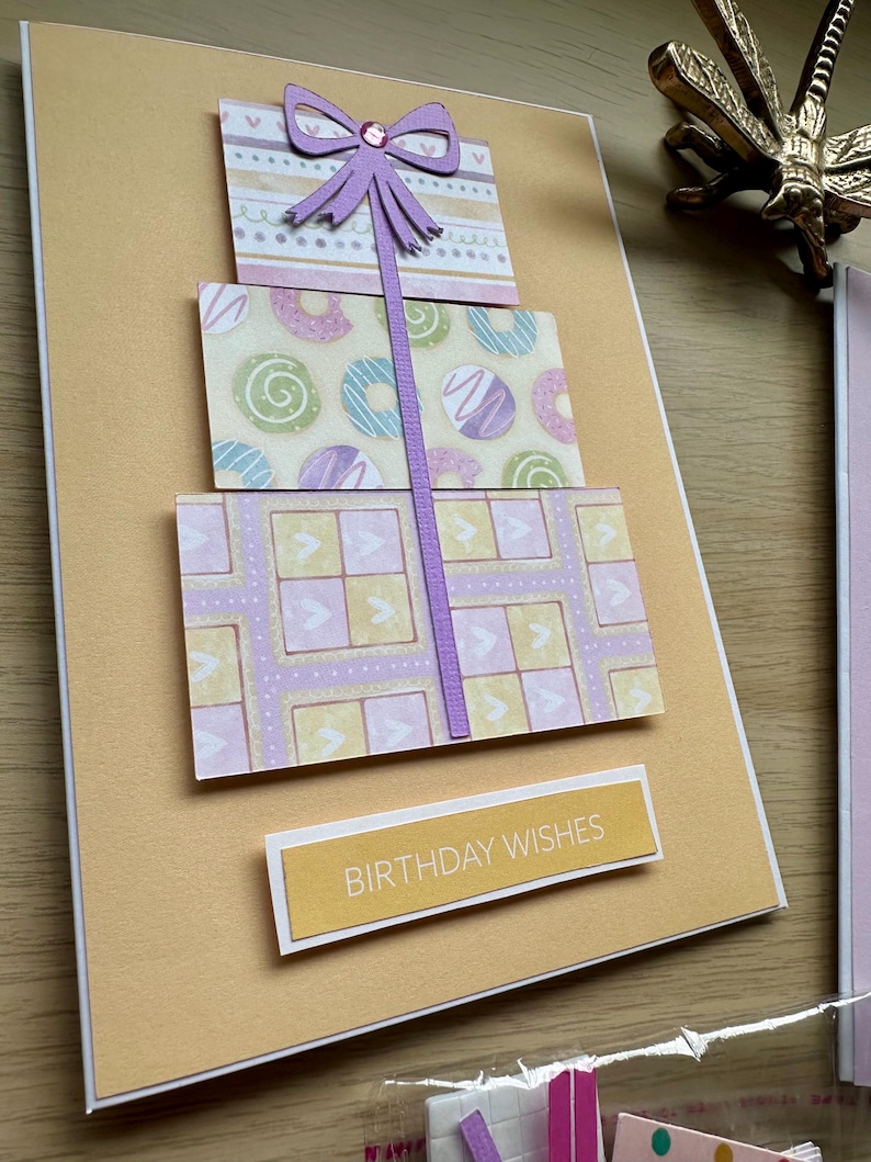 May include: A handmade birthday card featuring three stacked gift boxes with patterned paper. The top box has a purple bow. The middle box has donut designs. The bottom box has a geometric pattern. The card includes the text "BIRTHDAY WISHES".
