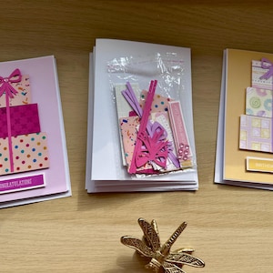 May include: Three handmade greeting cards with gift box designs. One card says "Congratulations" and another says "Birthday Wishes". A clear bag contains paper cutouts and embellishments. A gold dragonfly figurine is in the foreground.