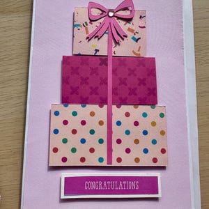 May include: A handmade greeting card with a tiered gift box design. The boxes are pink with patterned paper and a pink bow. The card includes the word "CONGRATULATIONS" on a rectangular label.
