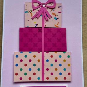May include: A handmade greeting card with a three-tiered gift box design. The boxes are pink and decorated with confetti, floral patterns, and polka dots. A pink bow and the word "Congratulations" complete the design.