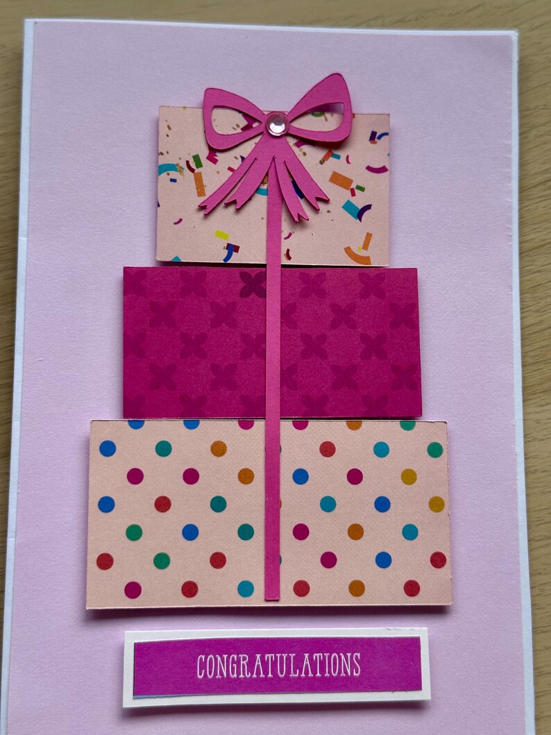 May include: A handmade greeting card with a three-tiered gift box design. The boxes are pink and decorated with confetti, floral patterns, and polka dots. A pink bow and the word "Congratulations" complete the design.