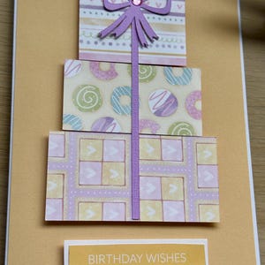 May include: A handmade birthday card featuring three stacked gift boxes in pastel colors. The top box has a purple bow with a gem, the middle box has a donut pattern, and the bottom box has a geometric design. The card includes the text "BIRTHDAY WISHES".