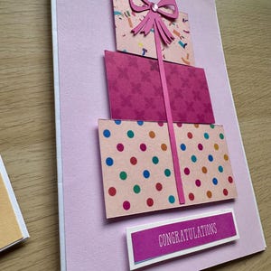 May include: A handmade greeting card with a pink background features three stacked gift boxes. The boxes have patterned designs in pink, with a pink bow and a ribbon. The bottom box has colorful polka dots. The card includes the word "Congratulations".