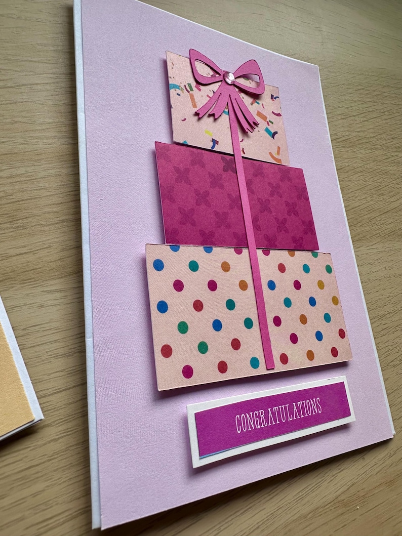 May include: A handmade greeting card with a pink background features three stacked gift boxes. The boxes have patterned designs in pink, with a pink bow and a ribbon. The bottom box has colorful polka dots. The card includes the word "Congratulations".