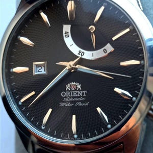 Op de afbeelding: Een close-up van een zilver- en goudkleurig Orient automatisch polshorloge. Het horloge heeft een zwarte, getextureerde wijzerplaat met goudkleurige wijzers en markeringen. De datum wordt weergegeven op 9 uur. De horlogeband is zilver- en goudkleurig.