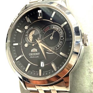 Könnte beinhalten: Silberfarbene Orient Automatik-Armbanduhr mit schwarzem Zifferblatt. Die Uhr verfügt über eine Tages- und Datumsanzeige, eine Mondphasenanzeige und ein silbernes Gliederarmband. Das Zifferblatt zeigt das Orient-Logo und den Text "Automatic".