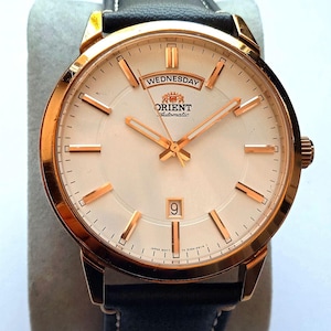 May include: A rose gold Orient Automatic watch with a cream-colored face, featuring gold hands and markers. The watch displays "Wednesday" and the date. It has a black leather band with white stitching.