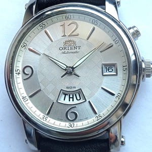 Könnte beinhalten: Silberfarbene Orient Automatik-Uhr mit schwarzem Lederarmband. Das Zifferblatt hat ein strukturiertes silbernes Zifferblatt, mit Anzeige von Tag und Datum. Die Uhr hat ein Edelstahlgehäuse und ein schwarzes Lederarmband mit weißen Nähten.