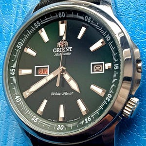 May include: A stainless steel Orient automatic watch with a dark green face, featuring white hour markers and hands. The watch displays the day and date, and is set on a black leather strap. The watch face has the text "Orient Automatic" and "Water Resist".